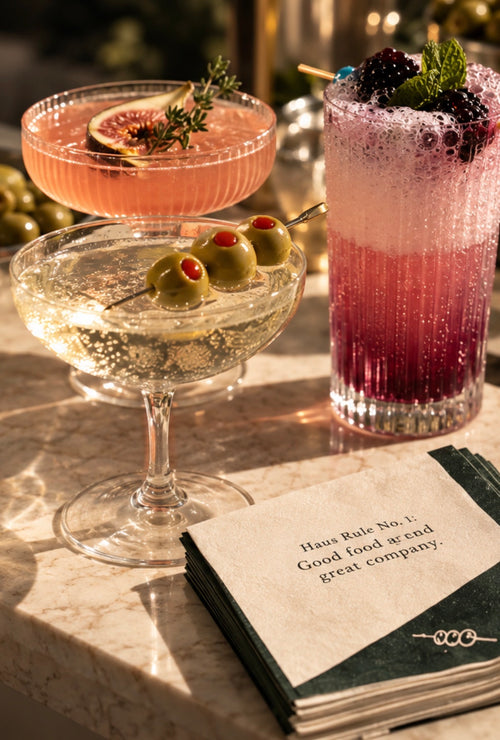 Three BarHaus drinks with fig, olive and blackberry garnishes beside a Haus Rule napkin