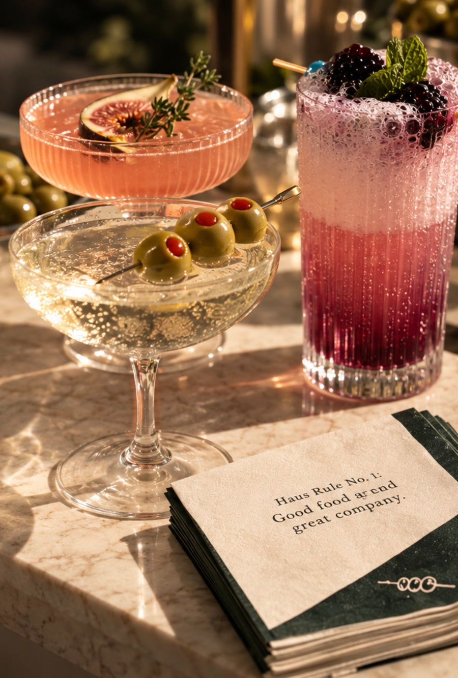 Three BarHaus drinks with fig, olive and blackberry garnishes beside a Haus Rule napkin