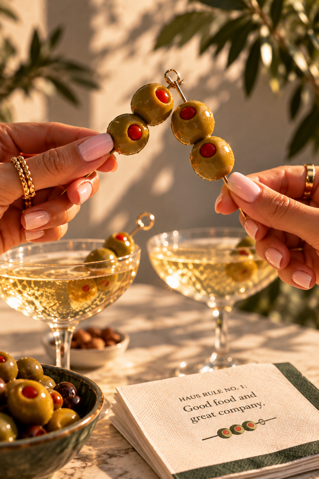 Two hands clinking olive skewers over coupe glasses beside a BiteHaus Haus Rule napkin
